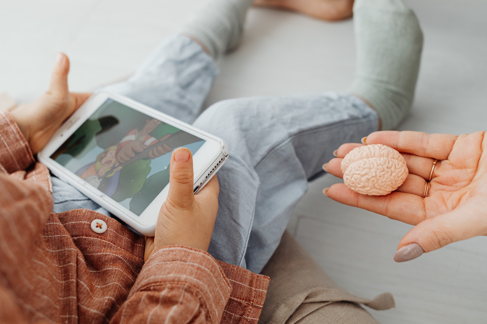 Kilkuletnie dziecko trzyma w rękach smartfon, rodzic wyciąga do niego otwartą dłoń na której leży przedmiot przypominający ludzki mózg.